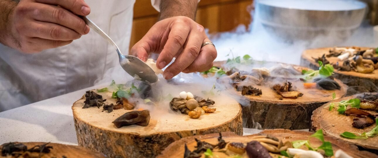 Flat lay of Alberta local foods including a dry-aged beef steak, fresh Saskatoon berries, a Caesar cocktail with celery salt rim, and Alberta honey on a wooden board