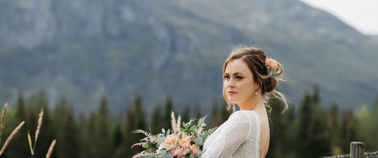 Bride in a lace wedding dress holding a peach and greenery bouquet stands by a wooden fence with Rocky Mountain views near Nordegg, Alberta.