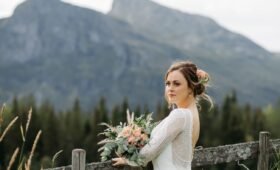 Bride in a lace wedding dress holding a peach and greenery bouquet stands by a wooden fence with Rocky Mountain views near Nordegg, Alberta.