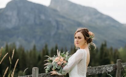Bride in a lace wedding dress holding a peach and greenery bouquet stands by a wooden fence with Rocky Mountain views near Nordegg, Alberta.