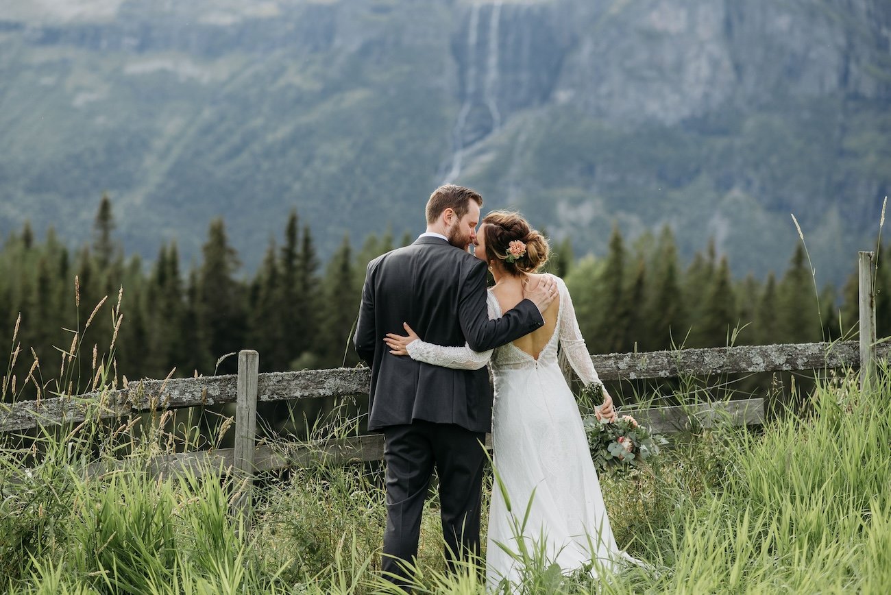 Bride and groom embracing by a wooden fence in tall grass with mountain and waterfall views during an outdoor wedding near Nordegg, Alberta.