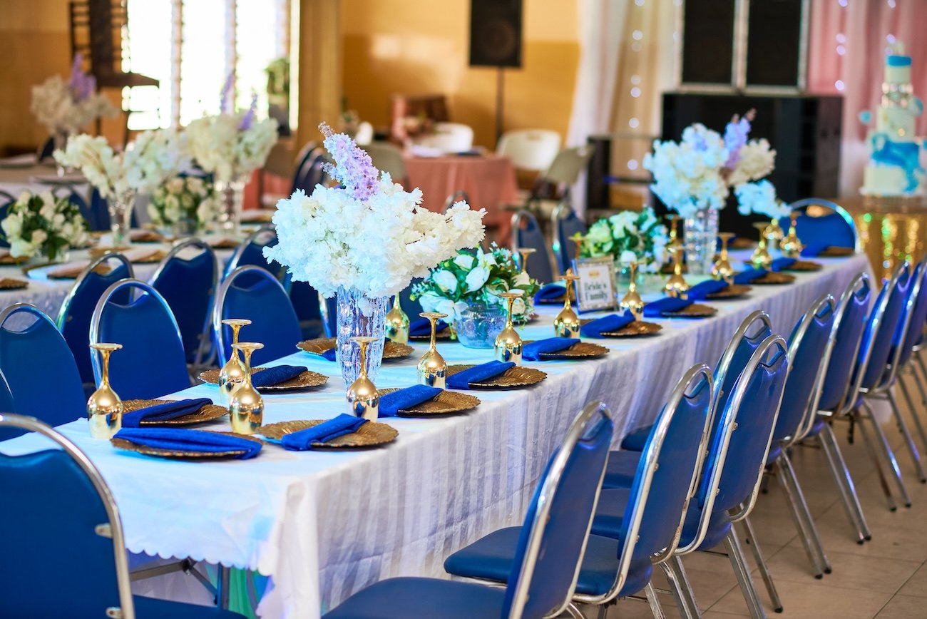 Long wedding reception table set with blue chairs, gold plates, and white floral centerpieces inside a hall near Nordegg, Alberta.